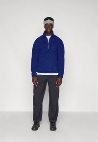 Maglione in pile blu con mezza zip, superficie strutturata, abbinato a pantaloni e scarpe nere, completato da un berretto grigio e occhiali da sole.