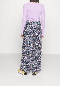 Kvinna som bär en lila långärmad topp, marinblå blommiga vida byxor och gula sneakers, stående mot en helt vit bakgrund.