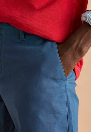Blaue Baumwollshorts mit glatter Textur und Seitentaschen, kombiniert mit einem roten kurzärmeligen Hemd, das ein lässiges Outfit zeigt.