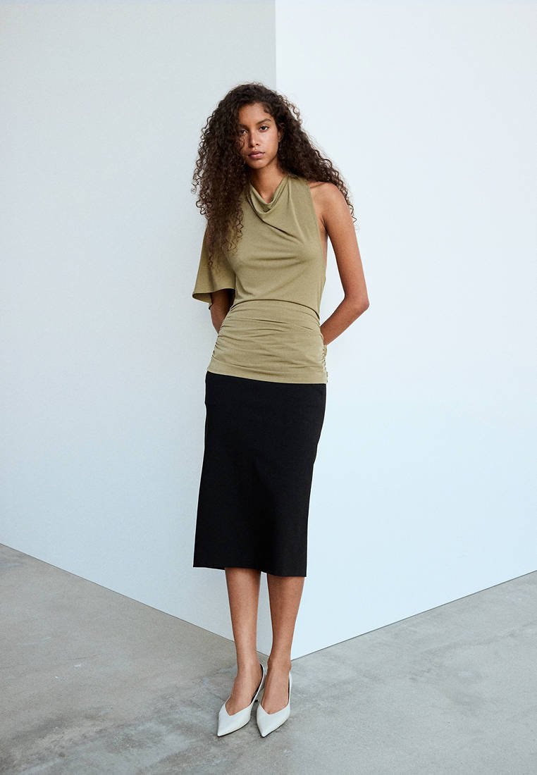 Jeune femme aux longs cheveux bouclés portant un haut asymétrique vert olive, une jupe noire longueur genoux et des talons blancs pointus, debout à l'intérieur.