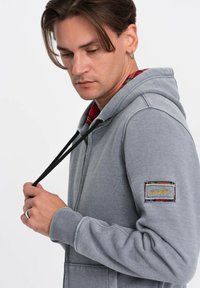 Felpa grigia con cappuccio e lacci neri, che presenta un patch testurizzato sul maniche sinistro e una fodera a quadri rossi visibile nel colletto.