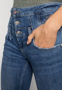 Jeans de ganga em azul médio, com uma cintura alta, três botões metálicos, bordas de bolsos desgastadas e um corte ajustado.