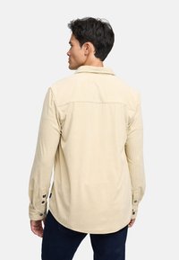 Camisa de pana beige con puños abotonados, un cuello con punta y un diseño de yugo en la parte posterior. Textura suave con surcos verticales.