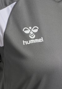 Tissu sportif gris et blanc avec un logo hummel blanc représentant une abeille stylisée et des chevrons sur la zone de la poitrine.