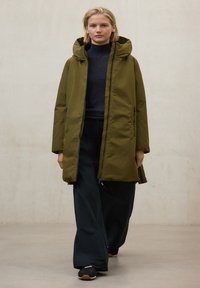 Parka verde oliva con capucha, cierre de cremallera y dos bolsillos laterales. Usada sobre un suéter oscuro y pantalones negros de corte holgado. Tejido texturizado.