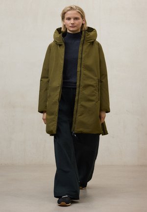 Parka verde oliva con capucha, cierre de cremallera y dos bolsillos laterales. Usada sobre un suéter oscuro y pantalones negros de corte holgado. Tejido texturizado.
