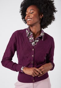 Lila stickad cardigan med rutförmönster, V-hals, knäppning och ribbad nederkant; lagd över en blommig skjorta med krage.