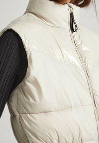 Jasnobeżowa puchowa kamizelka z wysokim kołnierzem, błyszczącymi panelami i zamkiem błyskawicznym z przodu. Teksturowany materiał po bokach dodaje kontrastu.