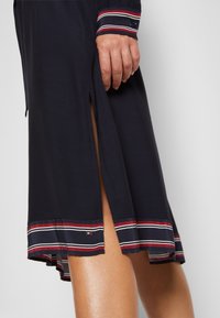Gros plan d'une robe bleu marine avec des poignets et un ourlet rayés rouge, blanc et noir, avec une fente latérale dévoilant une jambe nue en dessous.