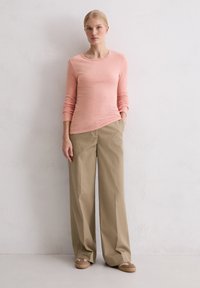 Langärmliges, hellrosa Hemd mit abgerundetem Saum, kombiniert mit beigen, weit geschnittenen Hosen. Das Outfit hat eine lässige, lockere Passform.