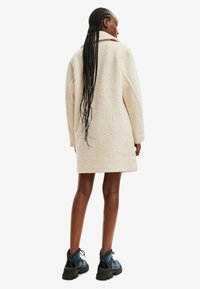 Kreemikas värvitooniga üleliigne mantel tekstuuriga kangast, millel on krae ja allavajutatud õlad, koos mustade ja siniste massiivsete saapatega.
