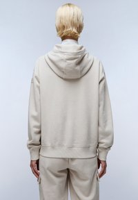 Beige oversized hoodie met een trekkoord capuchon, ribgebreide manchetten en onderkant. Gladde katoenen textuur. Draagt samen met bijpassende beige sweatpants.