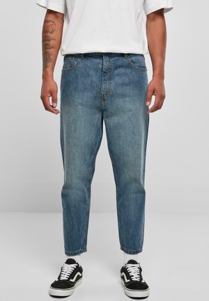 Homme portant un jean bleu moyen coupe décontractée, des chaussettes blanches, des baskets noires et blanches, et un t-shirt blanc avec les bras tatoués visibles.