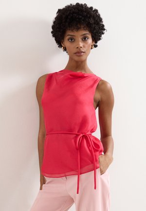 Femme aux cheveux courts et bouclés portant un chemisier rouge sans manches avec un nœud sur le côté, associée à un pantalon rose clair, debout contre un mur blanc.
