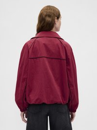 Jachetă roșie ușoară cu guler mandarin, manșete elastice și detaliu pe partea din spate, afișată din spate pe un fundal neutru.