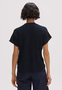 Persoon met kort, golvend donker haar, gekleed in een textuur zwart T-shirt met korte mouwen en donkere broek, staande met de rug naar de camera op een effen achtergrond.