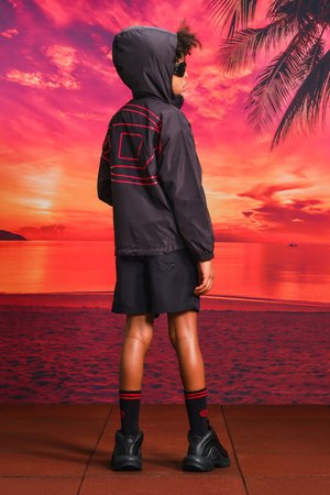 Giacca a vento nera con accenti rossi e tasche, abbinata a pantaloni corti neri, sneakers nere e calze a strisce rosse. Lo sfondo presenta un tramonto.
