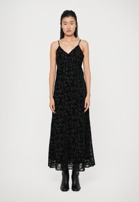 Abito maxi nero con sopra in pizzo, caratterizzato da una scollatura a V, spalline sottili e motivi floreali. Indossato con stivaletti neri alla caviglia.