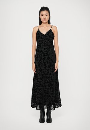 Abito maxi nero con sopra in pizzo, caratterizzato da una scollatura a V, spalline sottili e motivi floreali. Indossato con stivaletti neri alla caviglia.