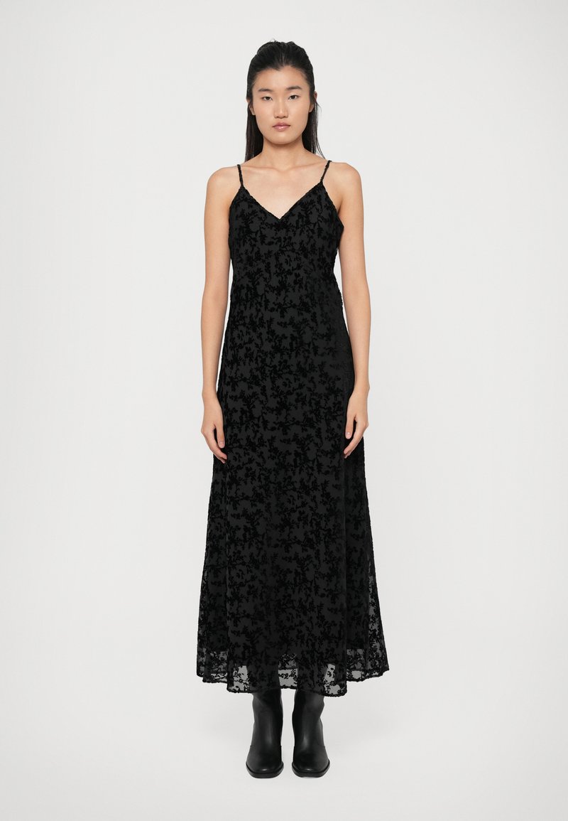 Abito maxi nero con sopra in pizzo, caratterizzato da una scollatura a V, spalline sottili e motivi floreali. Indossato con stivaletti neri alla caviglia.