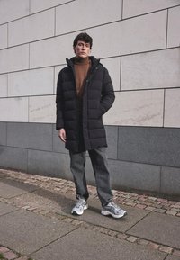 Cappotto imbottito nero con cappuccio sopra un dolcevita marrone, abbinato a jeans grigi larghi e sneaker grigie. Lo sfondo presenta pareti in pietra chiare e scure.