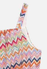 Primer plano de una correa de tela con patrón zigzag colorido y la parte superior del cuerpo de una prenda sin mangas sobre un fondo claro.