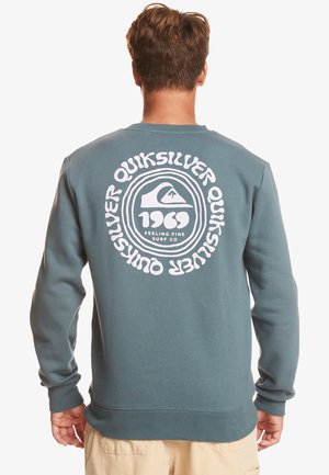Uomo che indossa una felpa blu Quiksilver con logo circolare bianco e testo "1969 Feeling Fine Surf Co" sul retro, di spalle, con pantaloncini beige visibili.