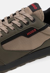 Zapatilla en caqui y negro con parte superior de malla texturizada, suela de goma y un destacado logotipo en rojo de "HUGO" en la lengüeta y el panel lateral.
