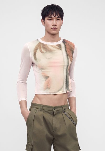 Νέος άνδρας που φοράει διαφανές crop top με μακριά μανίκια και μεγάλο τυπωμένο σχέδιο και φαρδιά ελιά πράσινα παντελόνια με αλυσίδα ως αξεσουάρ.
