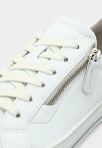 Gros plan d'une basket en cuir blanc avec des lacets crème et une fermeture éclair latérale argentée sur fond blanc.