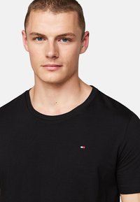 Svart crew neck t-skjorte laget av bomull, med et lite logo-patch i rødt, hvitt og blått på venstre bryst. Glatt tekstur.