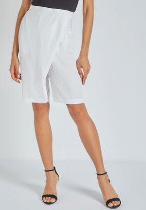 Pantalones cortos blancos hasta la rodilla con una tela texturada, que presentan una cinturilla lisa y un sutil diseño con pliegues. Combinados con sandalias negras de tiras.