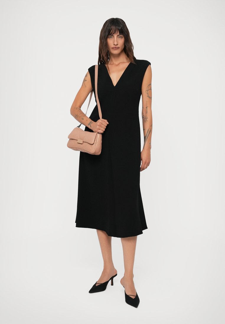 Femme avec des cheveux châtain mi-longs portant une robe noire sans manches, des talons noirs et un sac à bandoulière beige, se tenant debout avec des tatouages sur les bras.