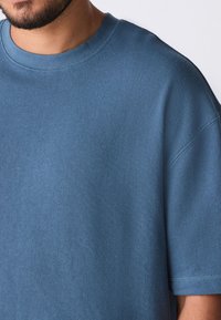 Camicia corta a maniche corte in cotone blu con collo rotondo, dotata di un tessuto testurizzato e vestibilità ampia. Le cuciture sono visibili attorno al collo e alle maniche.
