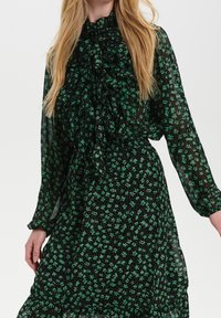 Svart klänning i genomskinligt tyg, med ett grönt blommönster, volangkrage och långa, puffärmar. Elastisk midjedetalj.