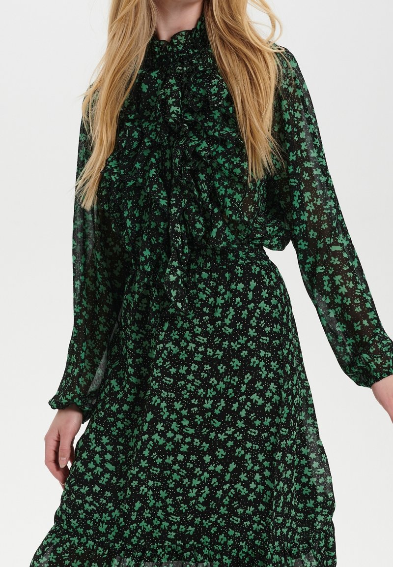Svart klänning i genomskinligt tyg, med ett grönt blommönster, volangkrage och långa, puffärmar. Elastisk midjedetalj.