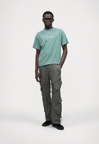 T-shirt verde de algodão com texto branco, emparelhado com calças de trabalho cinzentas com vários bolsos e punhos ajustáveis. Sapatos pretos completam o visual.
