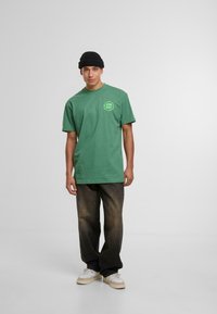 Camiseta de algodón verde con cuello redondo y un logo en el lado izquierdo del pecho, combinada con jeans de mezclilla oscuros y holgados y zapatillas blancas.