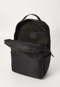 Sac à dos noir en tissu lisse, avec un dessus zippé, une poche avant et une étiquette à l'intérieur. Design minimaliste et forme structurée.