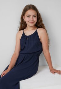 Navyblaues ärmelloses Kleid mit gerafftem Ausschnitt, taillierter Taille und fließendem Stoff. Das Model hat lange, wellige Haare und sitzt vor einem hellen Hintergrund.