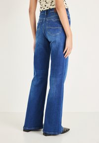 Kék bővülő szárú farmernadrág, magas derékkal, hátul zsebekkel és finom varrási részletekkel. A szárak fokozatosan szűkülnek a szegély felé.