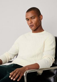 Suéter de punto trenzado crema con puños acanalados y cuello redondo, combinado con pantalones de pana verde oscuro. Modelo sentado en una silla de cromo.