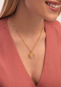 Goldene Anhängerkette an einer zarten Kette, mit einem strukturierten achteckigen Anhänger mit aufwendigen Designdetails, getragen über einem rosa Stoff.