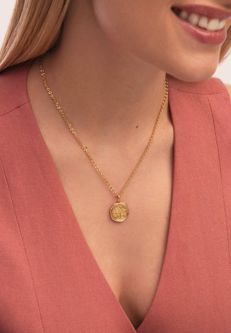 Goldene Anhängerkette an einer zarten Kette, mit einem strukturierten achteckigen Anhänger mit aufwendigen Designdetails, getragen über einem rosa Stoff.