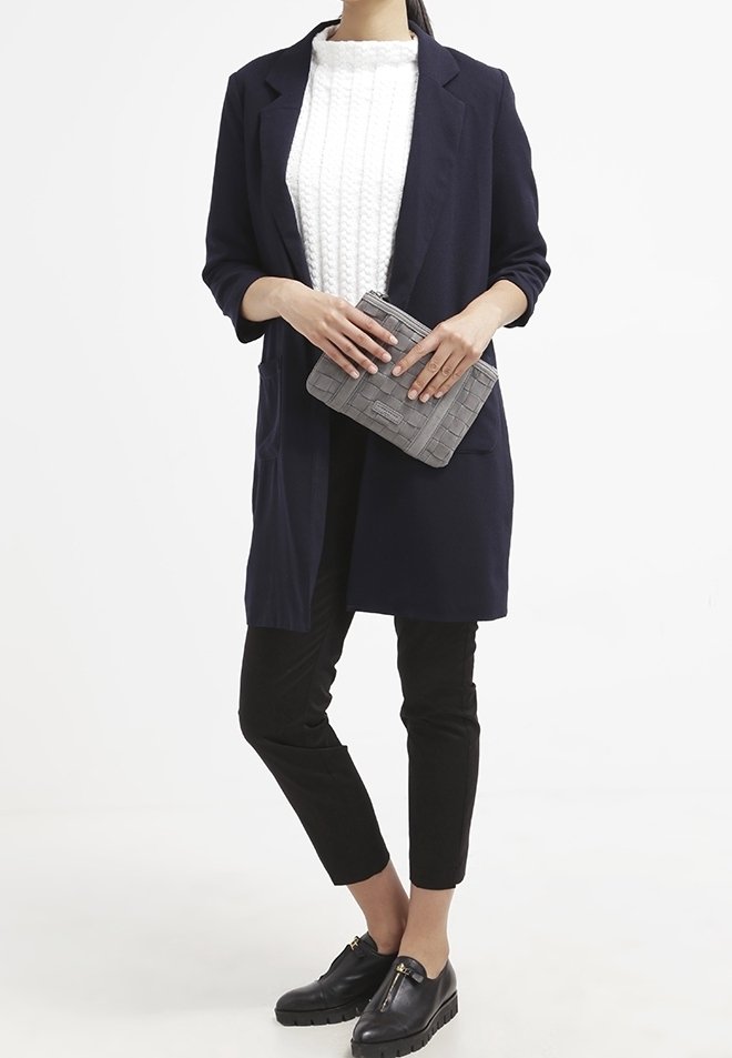 Femme portant un manteau bleu marine, un pull blanc texturé, un pantalon court noir, des mocassins noirs, tenant une pochette grise texturée.