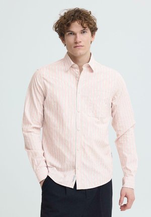 Ung mand med krøllet hår iført en lyserød og hvid stribet skjorte med knapper og sorte bukser, stående foran en ensfarvet baggrund.