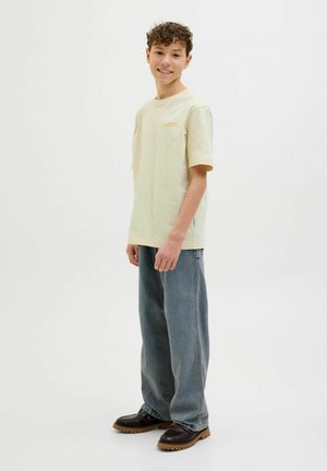 Tricou din bumbac de culoare galben deschis, cu mâneci scurte și guler rotund, asortat cu blugi din denim albastru, lejeri, și pantofi maro închis.