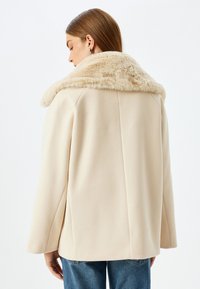 Beige ullblandad kappa med en mjuk krämfärgad pälskrage, skräddarsydd form, långa ärmar och en delning bak, stylad med blå jeans.