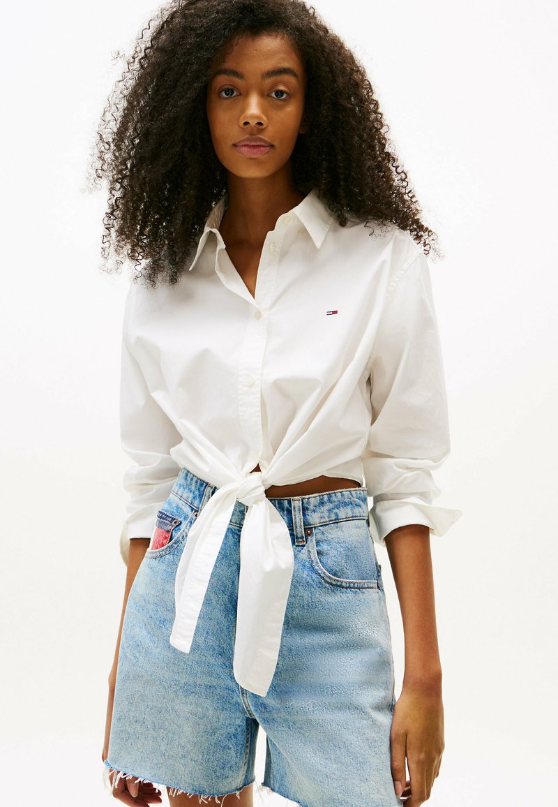 Camicia bianca in cotone con bottoni legata in vita, abbinata a pantaloni corti in denim blu chiaro con orlo sfilacciato e una piccola etichetta rossa sulla tasca.