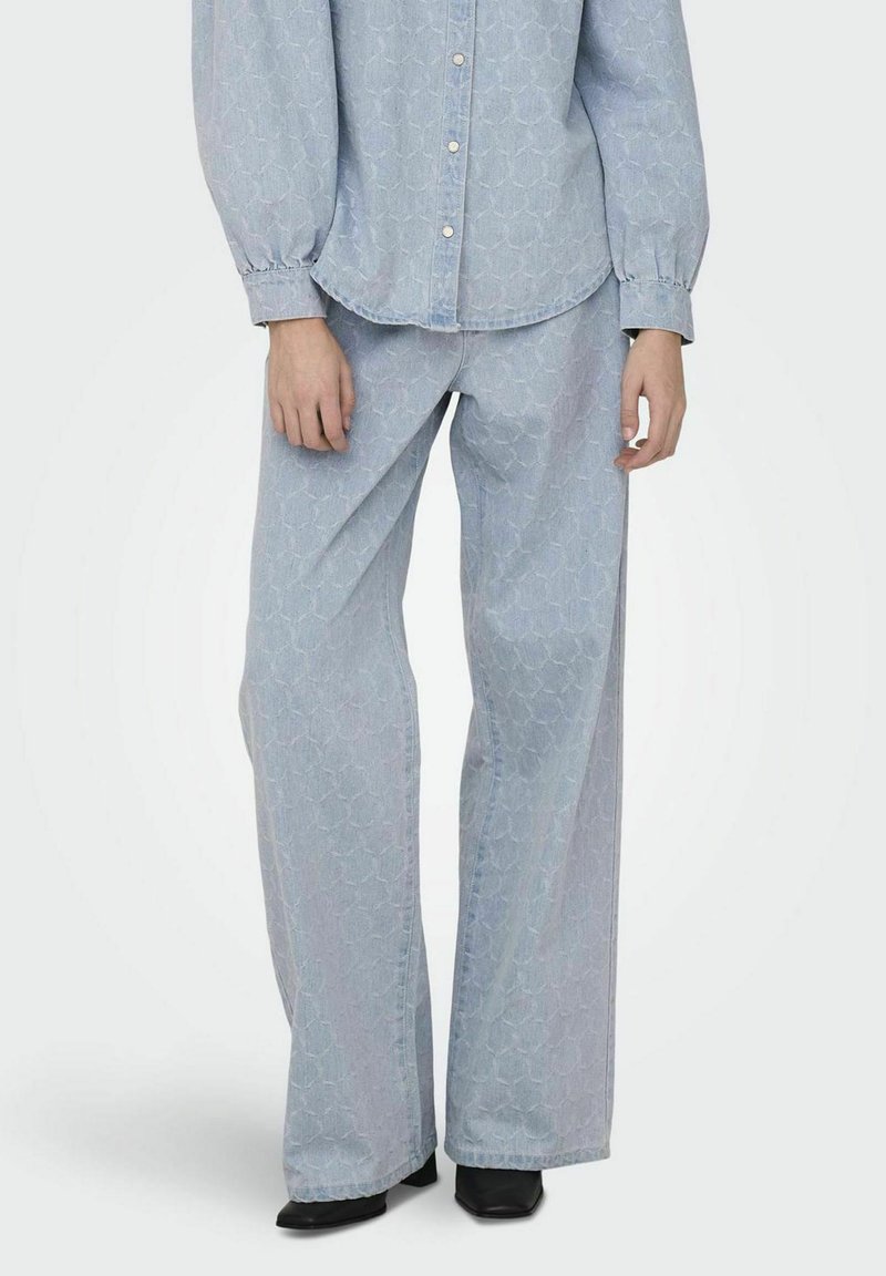 Lichtblauwe denimoutfit met een oversized gestreept shirt en wijde broek, aangevuld met een subtiele honingraat textuur en puntige zwarte schoenen.